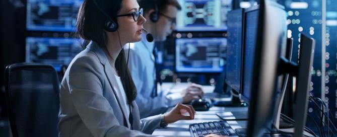 female police dispatcher with male colleague in background looking at computer screens